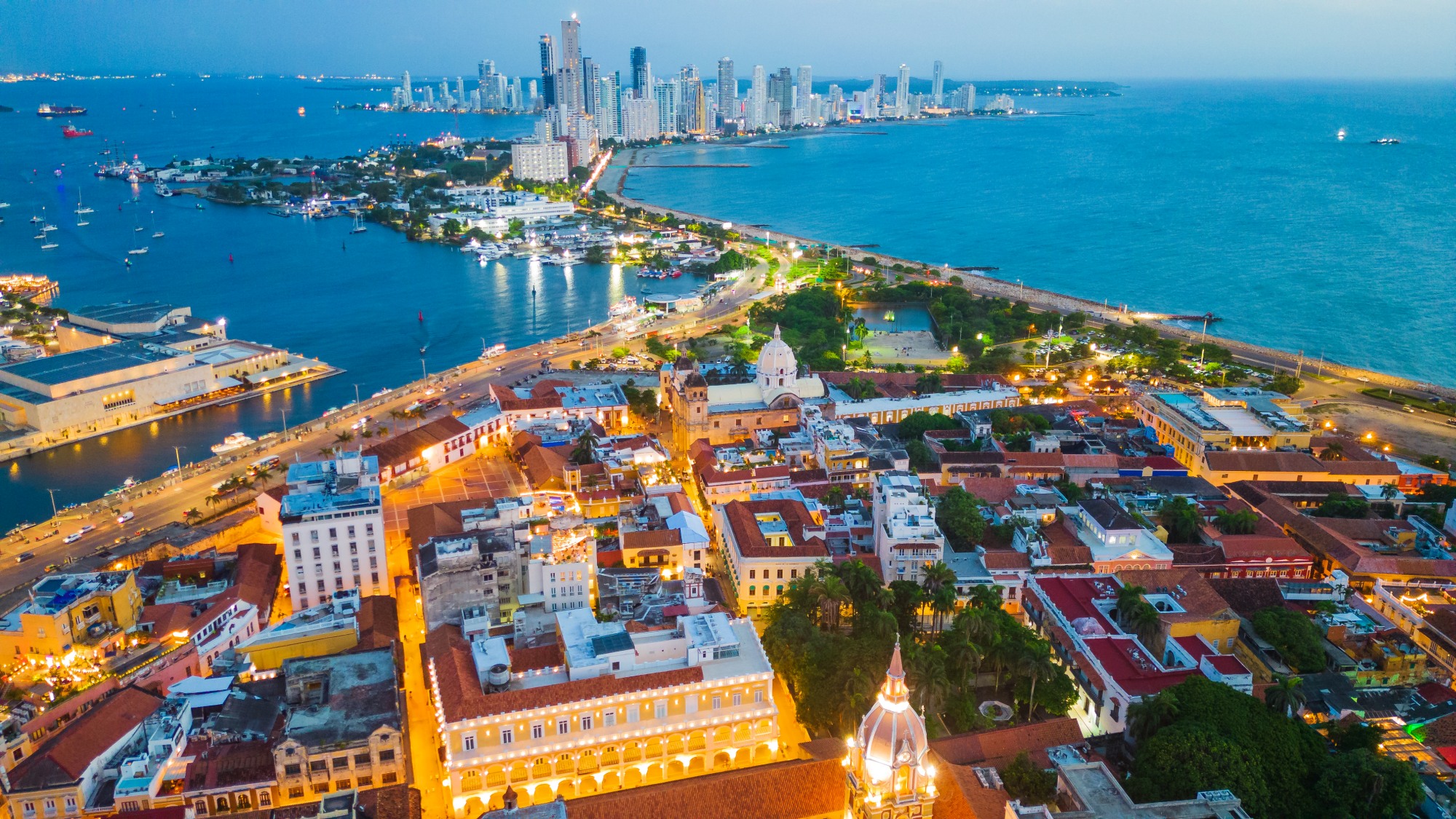 Aerial view of Cartagena at dusk: the lit old town in the foreground, the bay and modern skyline behind